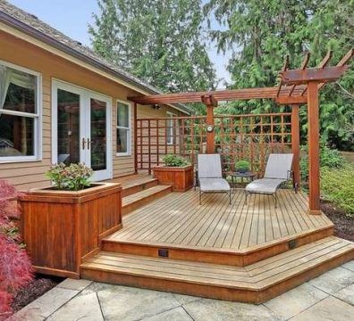 Wooden deck with steps, trellis, pergola, two chairs, and planter boxes next to a house.