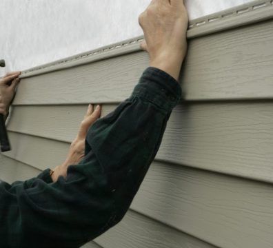 Person installing beige vinyl siding on a house, using a hammer and hands.