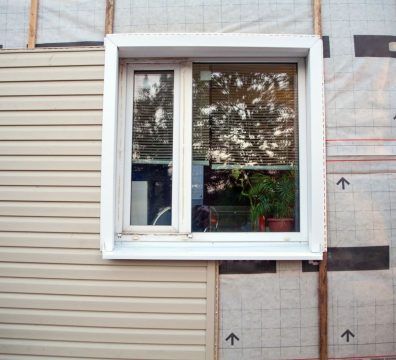 Window with white frame, tan siding, and exposed house wrap. Window has blinds and a potted plant.