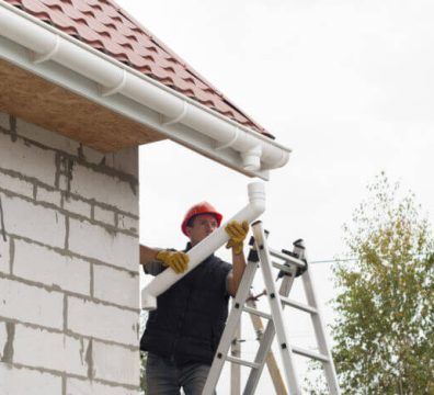 Man installing a white gutter downspout on a building with a red roof, standing on a ladder.