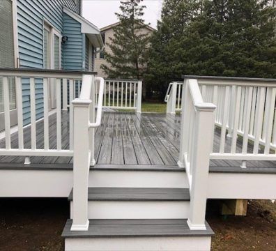 Gray and white deck with steps leading to a house, surrounded by railing and greenery.