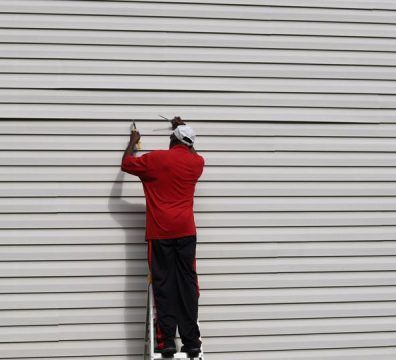 Person on a ladder, working on light gray vinyl siding; wearing red shirt and black pants.
