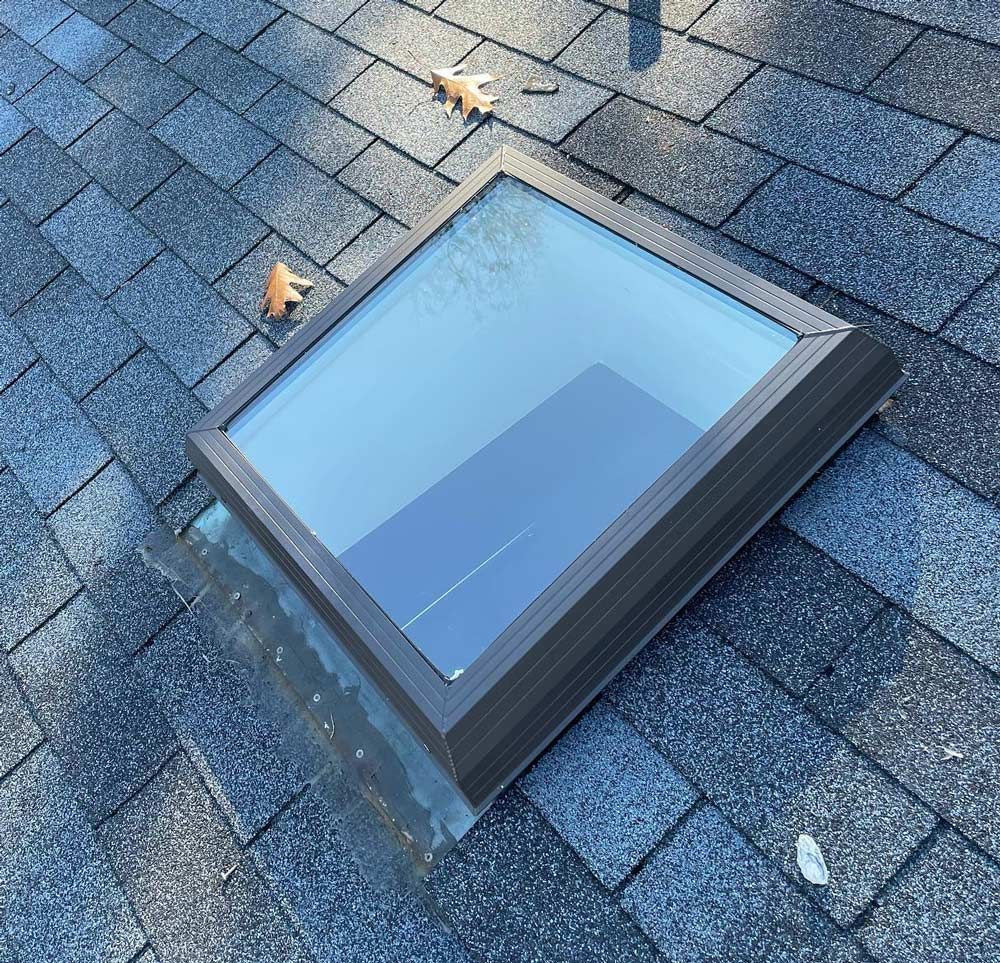 Skylight on a shingled roof, showing a clear view of the sky. Brown metal frame and surrounding shingles.