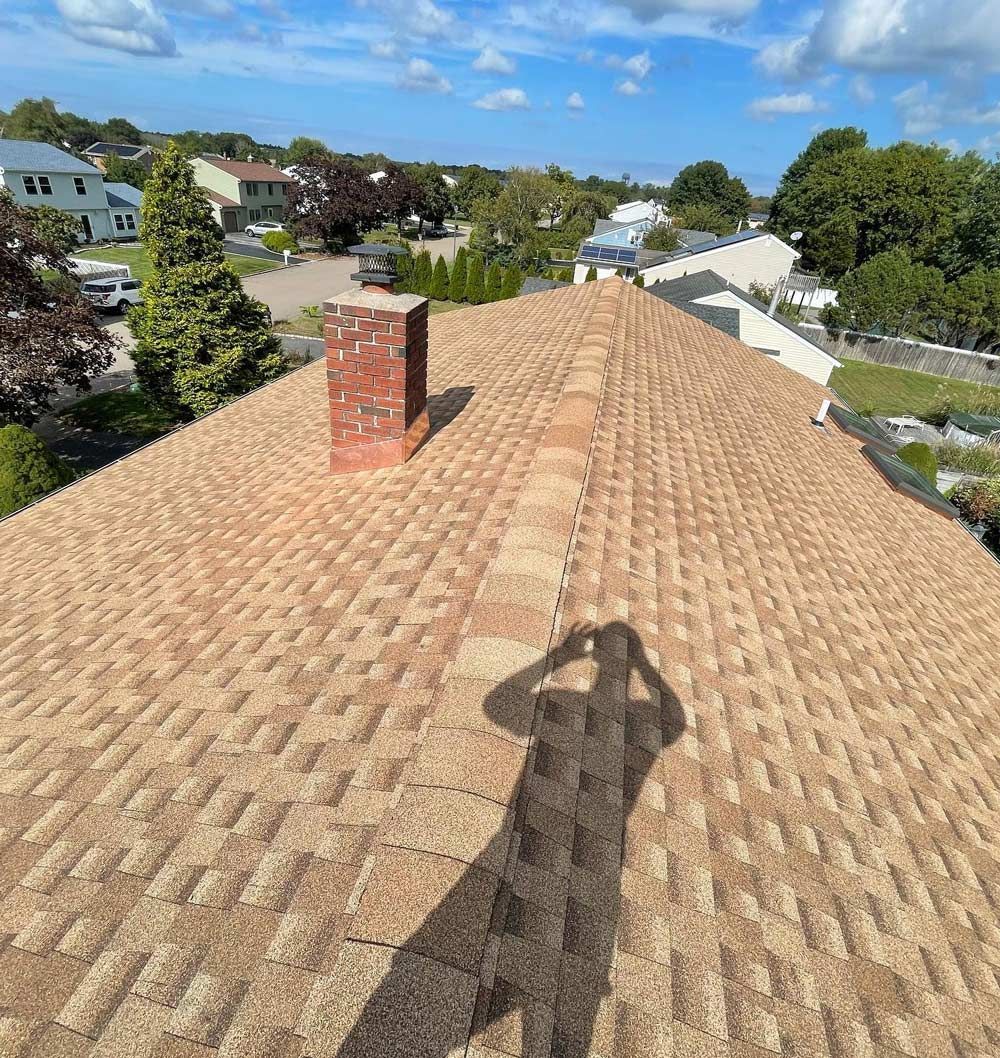 A person's shadow on a textured brown roof with a brick chimney under a blue sky.