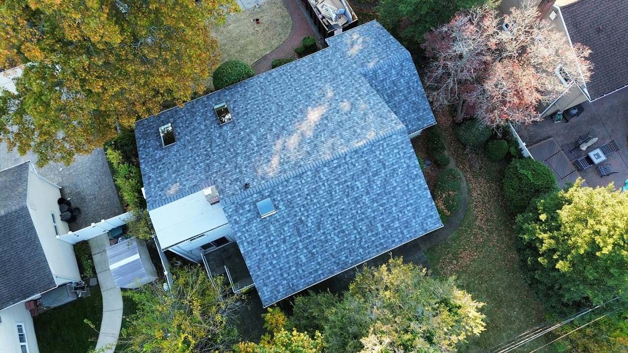 Overhead view of a house with a blue-gray shingled roof, surrounded by green and autumn-colored trees.