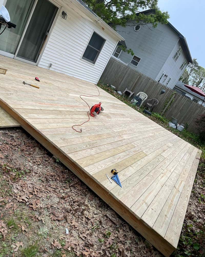 A partially built wooden deck with tools scattered on the surface, next to a white house with a slanted roof.
