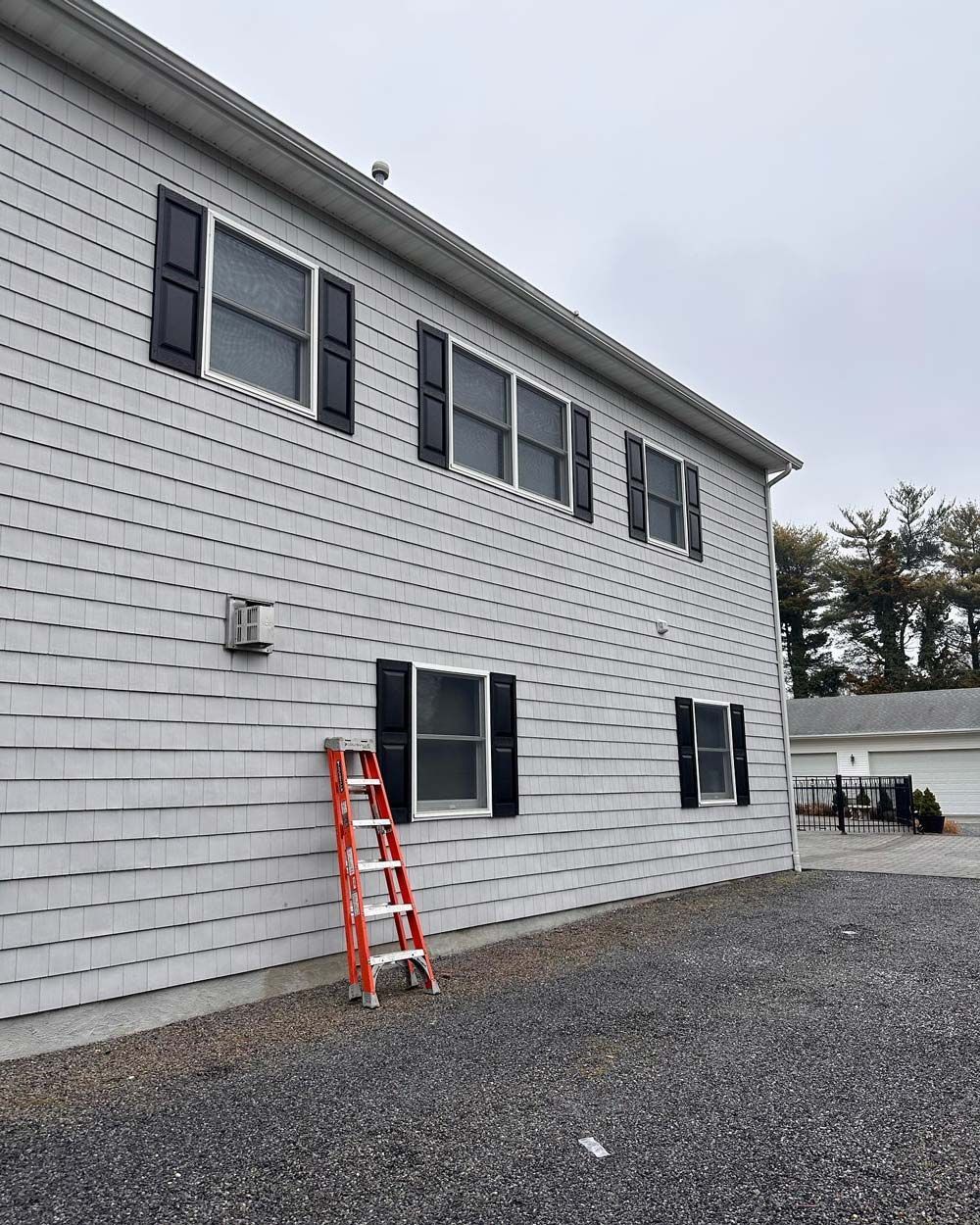 Gray house with black shutters; red ladder propped against the siding. Gravel driveway.