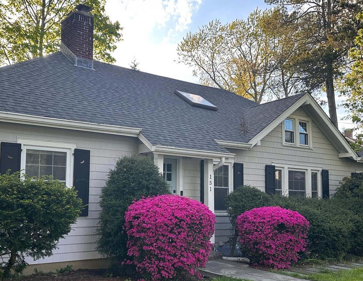 Cottage-style home with grey roof, black shutters, and blooming pink azaleas in front.