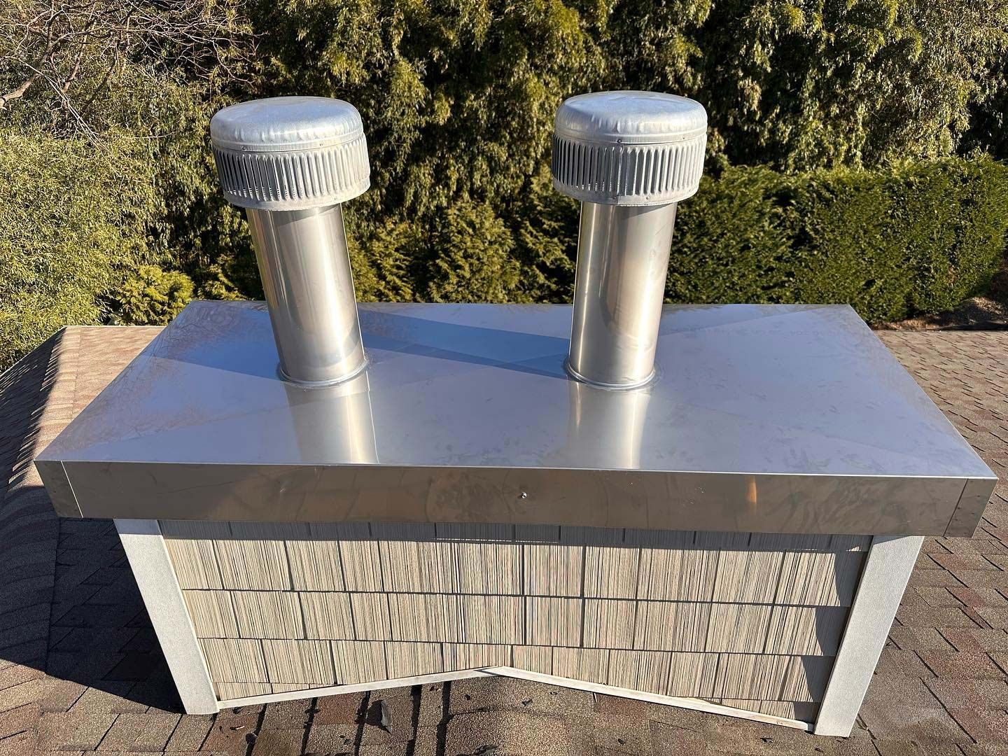 Metal rooftop vent with two silver exhaust pipes, with a decorative brick-like base, outdoors in sunlight.