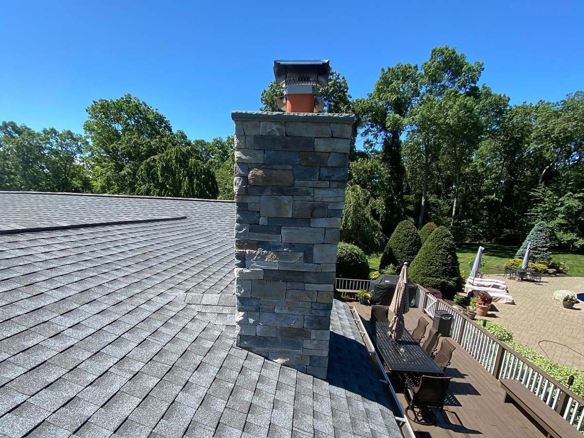 Stone chimney on a gray shingled roof, set against a bright blue sky and green trees.