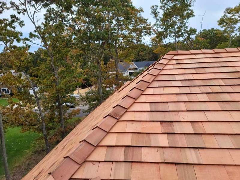 Close-up view of a new cedar shake roof with reddish-brown wood shingles, trees, and overcast sky in the background.