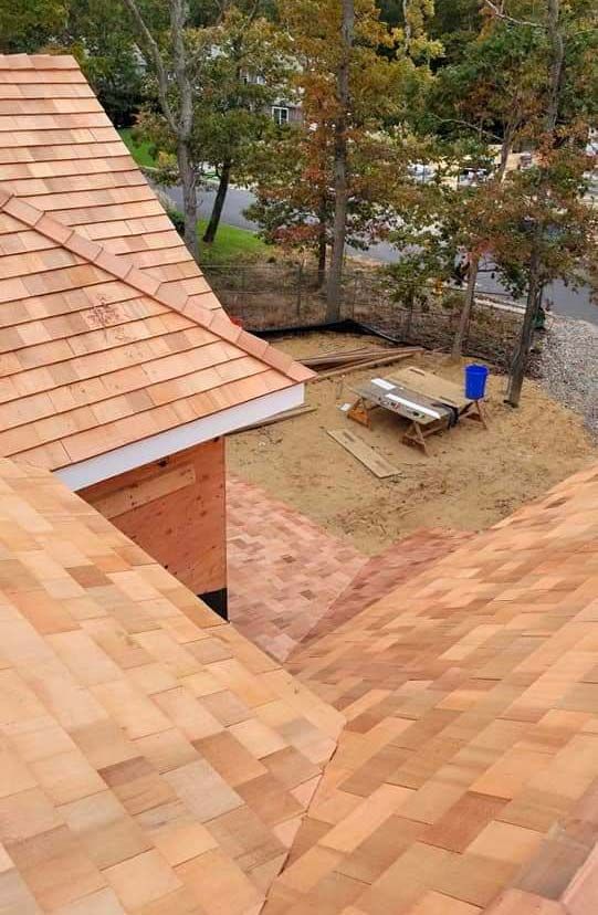 Wooden shingle roof under construction, surrounded by trees and a dirt yard.