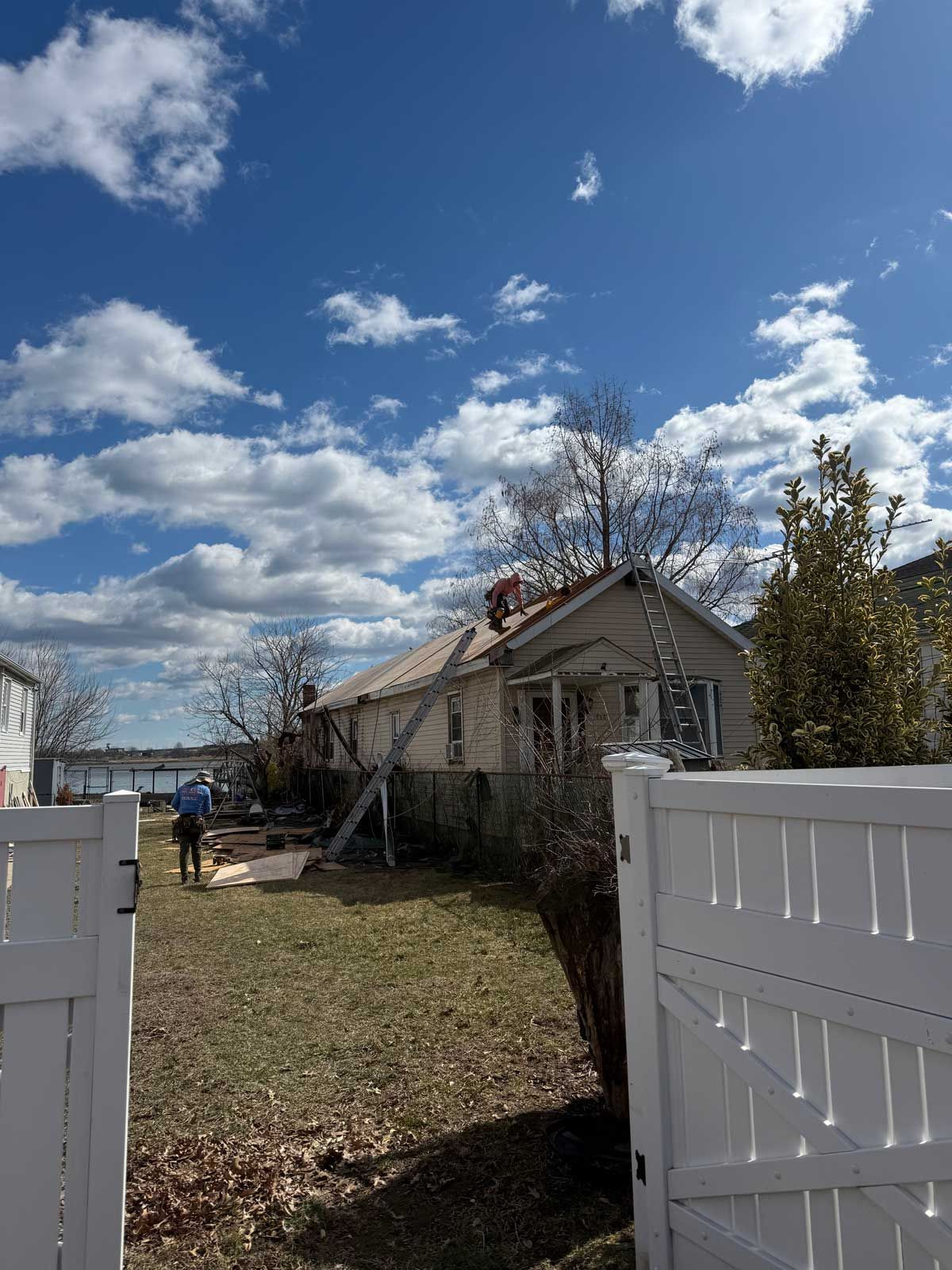 Fence frames a yard leading to a one-story house under a blue sky with fluffy clouds.