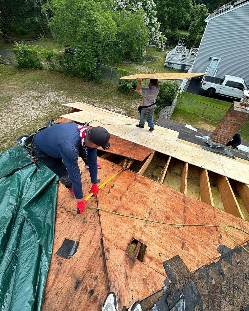 Two roofers replacing a damaged roof, one measuring with tape, the other carrying a plank. Outdoors, sunny.