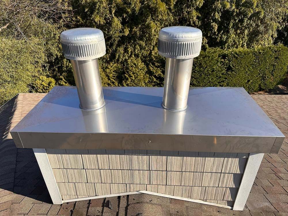 Stainless steel chimney cap with two vents, set outdoors, brick base, surrounded by greenery.