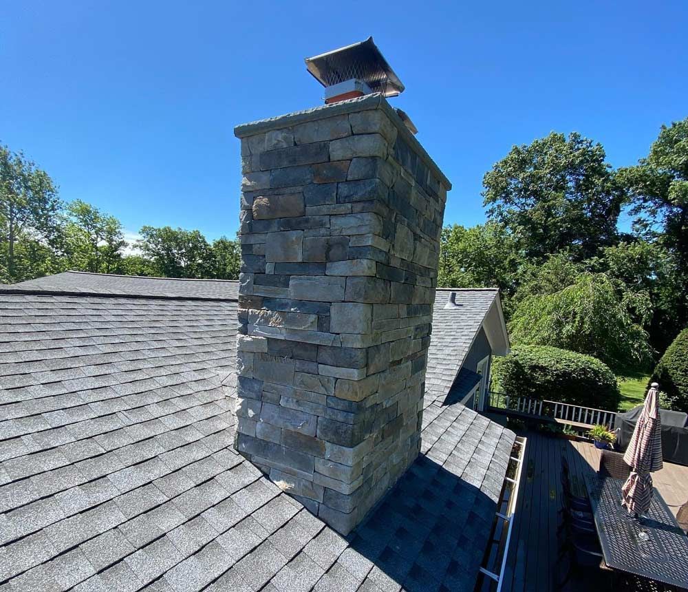 Stone chimney on a shingled roof against a bright blue sky, surrounded by green trees.