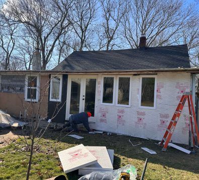 House undergoing renovations, siding installation in progress. Worker near new windows, ladder, insulation materials.