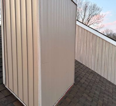 Chimney clad in tan siding, surrounded by matching siding on a roof with brown shingles.