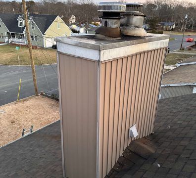 Chimney on rooftop, tan metal siding, three vents, in residential area.