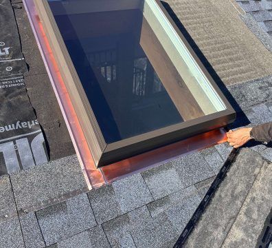 A rooftop skylight with copper flashing, partially installed, and a person's hand adjusting a shingle.