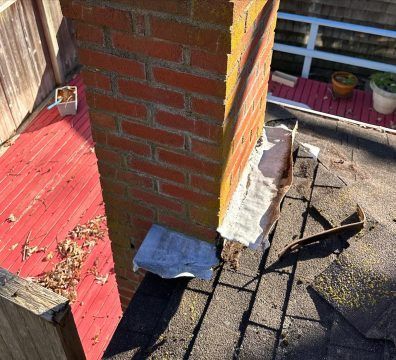 Brick chimney on a shingled roof with damaged flashing, red wooden deck, and leaves.