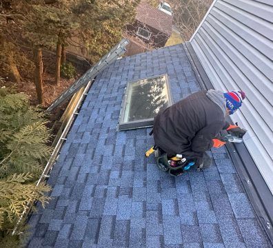 Roofer on a blue shingle roof, cutting shingles near a skylight, with a toolbox.