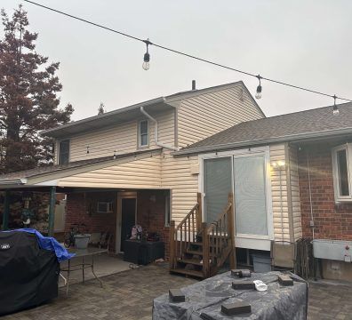 Backyard of a two-story house with string lights. A covered patio, brick patio and stairs are visible. Overcast sky.