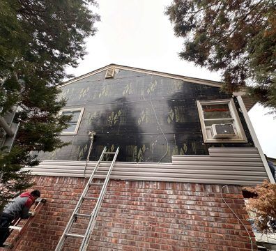 Person installing siding on a house. The house has exposed sheathing, brick, and installed siding. A ladder is present.