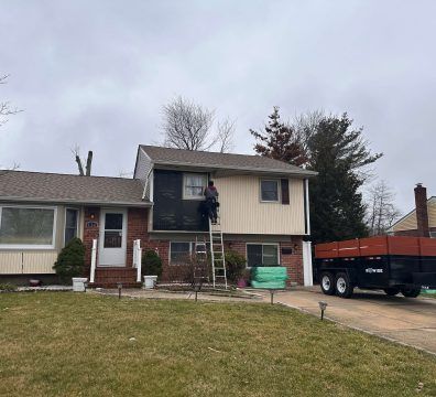 Person on ladder siding a two-story beige and brick house. Flatbed truck in the driveway.