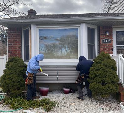 Two workers installing siding on a house with a bay window. Grey siding and white trim.