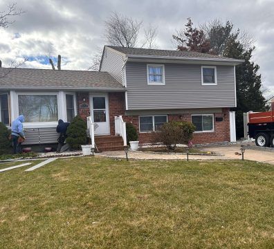 House exterior with brick and gray siding. Workers repair siding on a cloudy day.