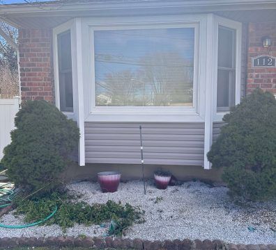 Bay window of a brick house with bushes, gravel, and two flower pots. Beige siding below the window.