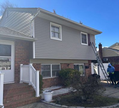 Two-story house with gray siding; a ladder leads to the roof, person is on the ladder.