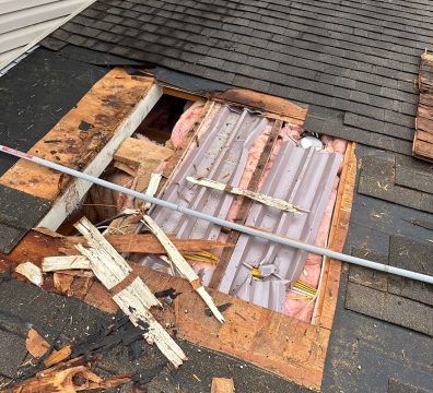 Damaged roof with hole, exposed insulation, wood, and metal, likely from storm damage.