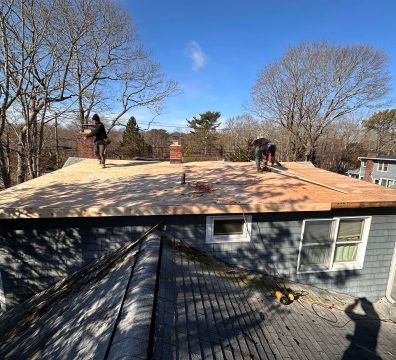 Roofers installing new roofing on a home, visible are plywood and tools on the roof under a blue sky.