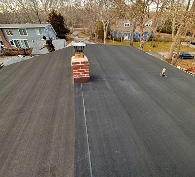 Black flat roof with a brick chimney. A person working on the roof with houses in the background.