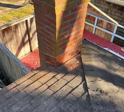 Brick chimney with copper flashing on a roof, surrounded by shingles and wooden fence.