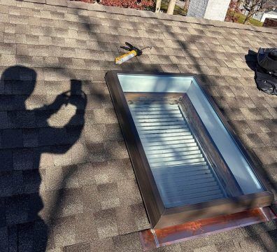 Person's shadow taking photo of a skylight on a shingled roof with a caulk gun.