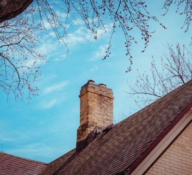 Brick chimney atop a brown shingled roof against a bright blue sky with tree branches.