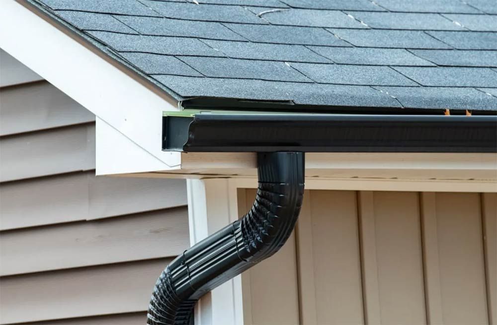 Black gutter and downspout attached to a roof and beige siding.