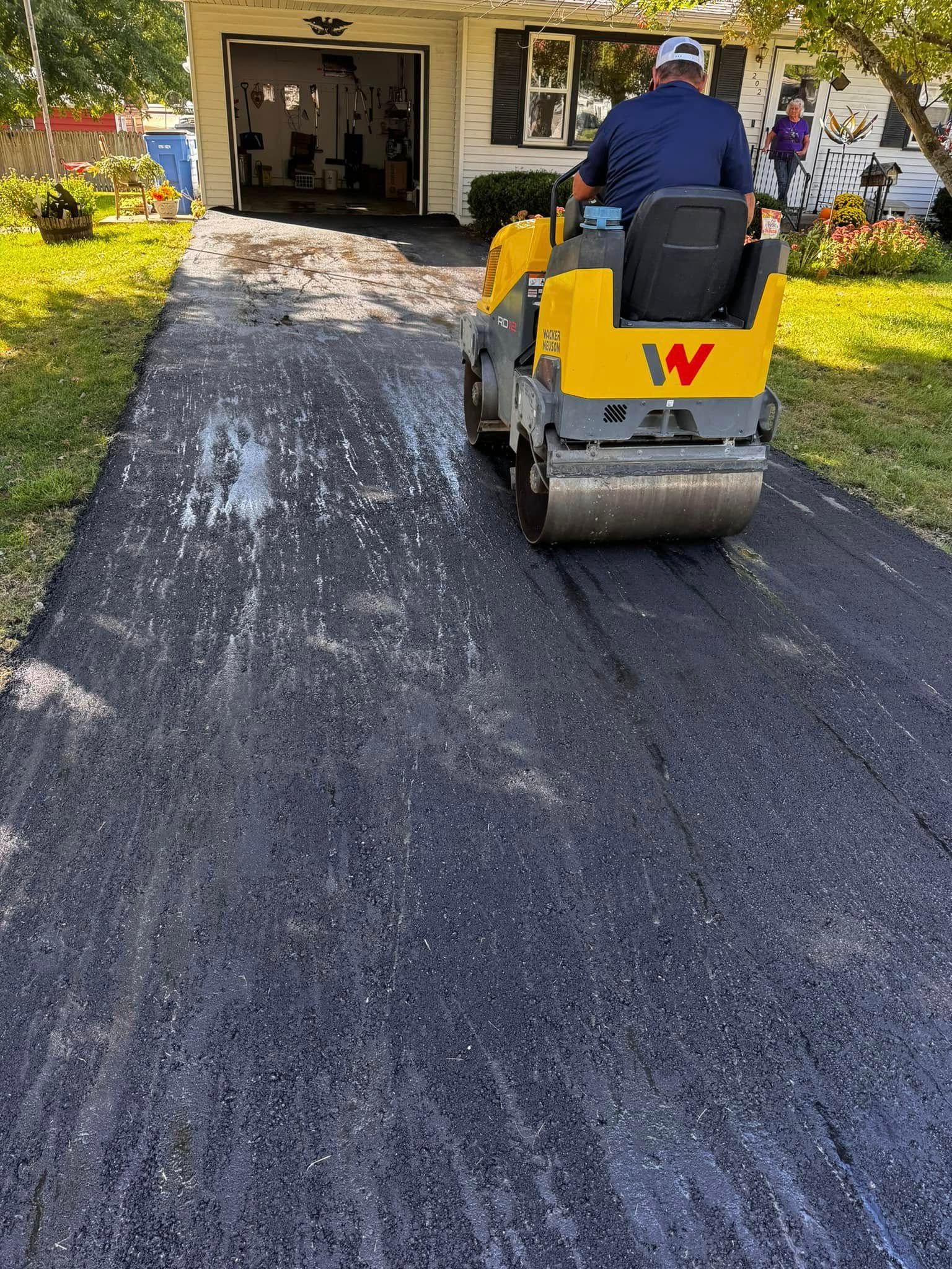 Asphalt roller compacting a fresh driveway; yellow machine, person driving.
