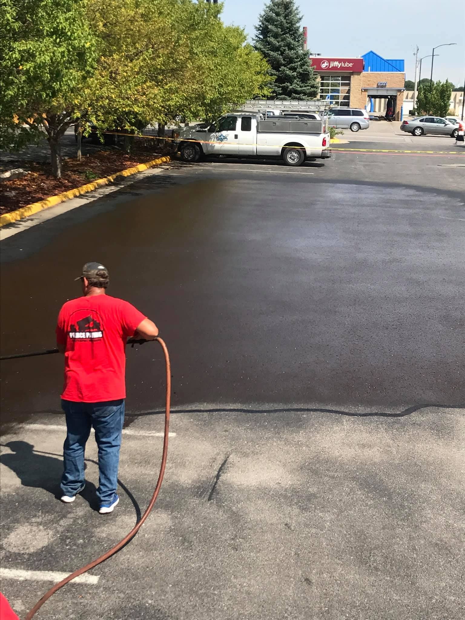 Man spraying sealant on a parking lot. A white truck and building are in the background.