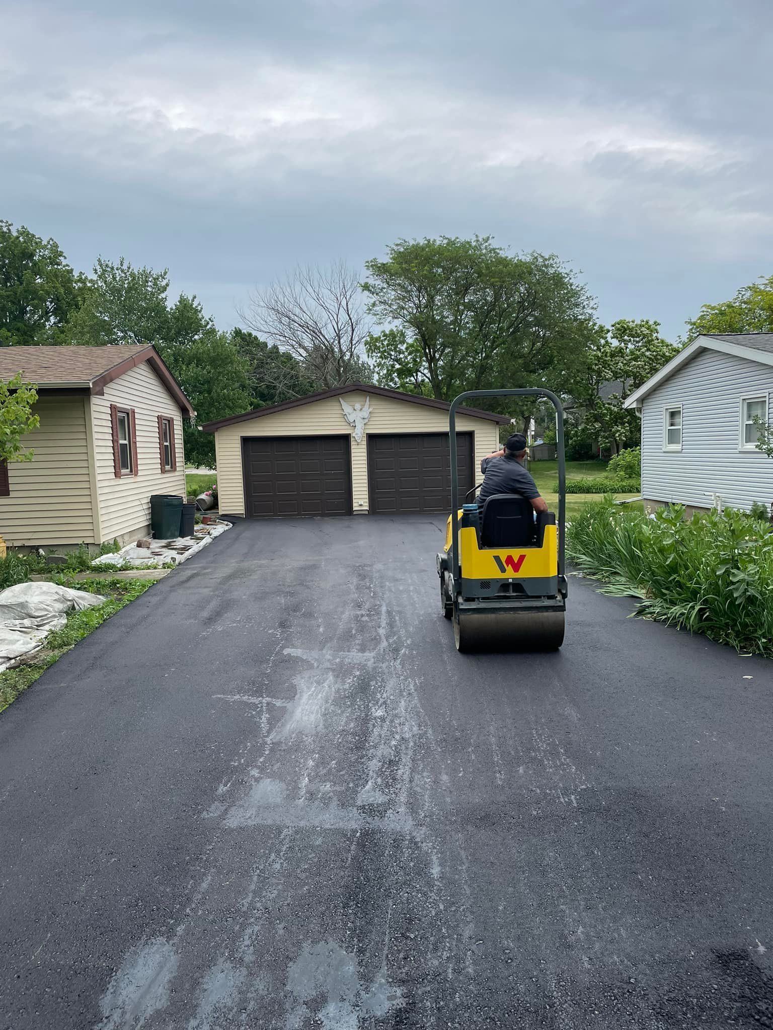 Person compacting fresh asphalt on a driveway with a roller in front of a garage, under a cloudy sky.