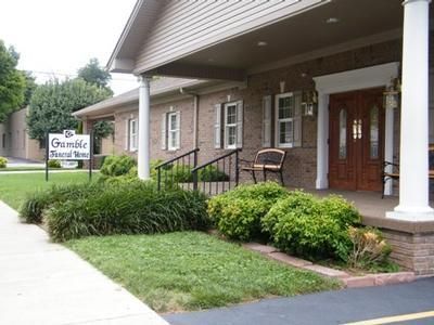 Covered entrance of a brick building with white columns, a bench, shrubs, and a sidewalk sign.