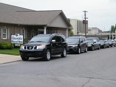 SUVs and cars lined up along a street beside a small building with a sign, under overcast skies