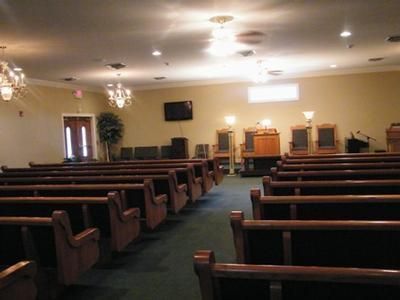 Empty church sanctuary with rows of wooden pews, ceiling lights, and a wall-mounted TV at the front