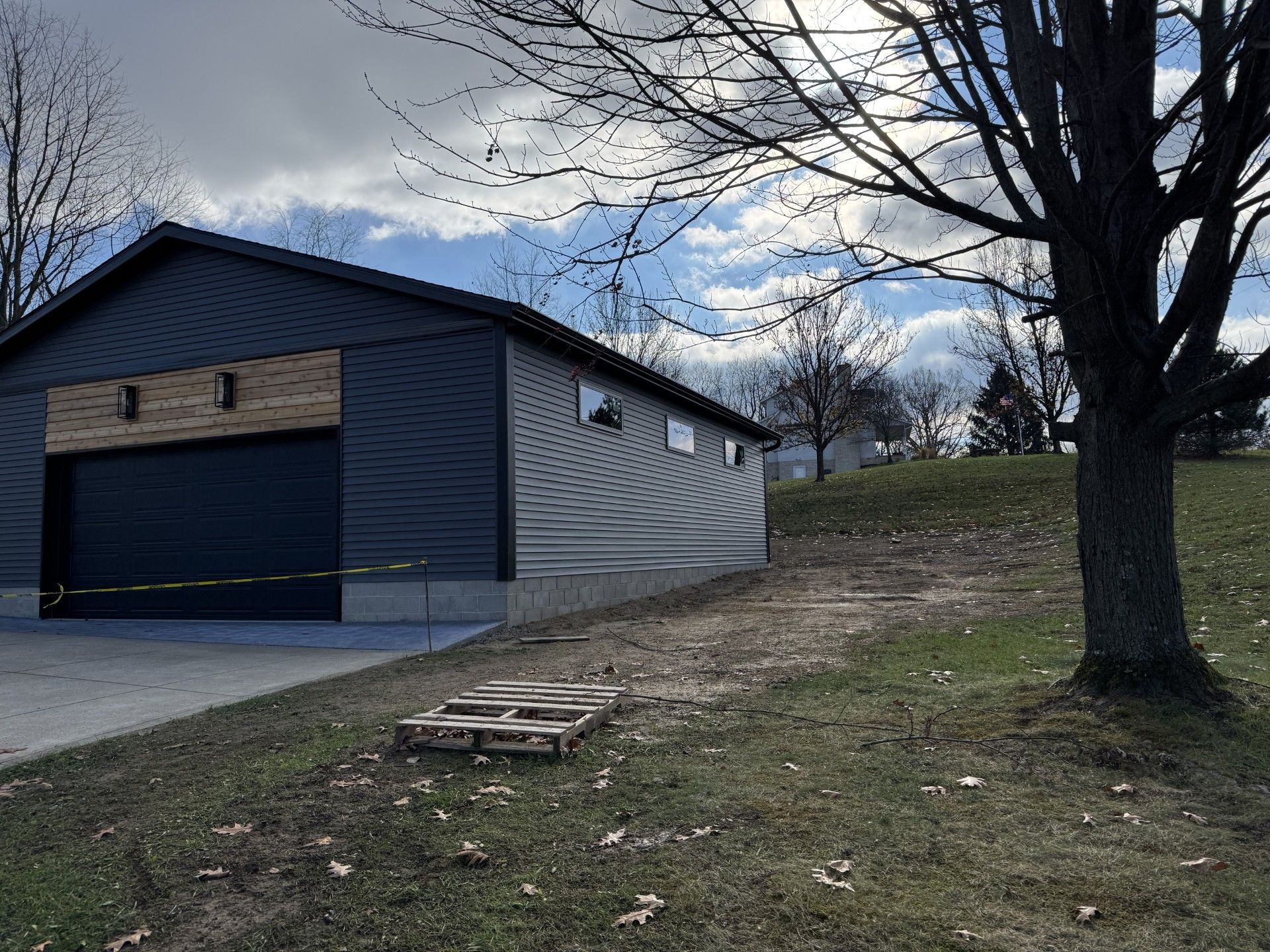 Blue-sided garage or shed beside a driveway, with bare trees and a grassy yard under a cloudy sky.