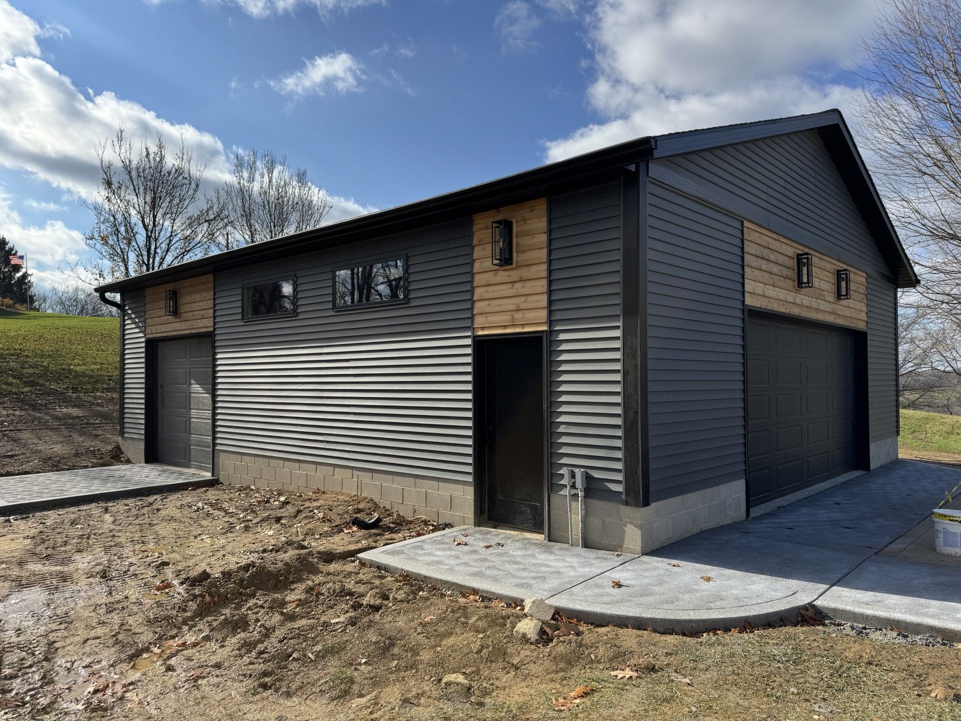 Modern gray metal garage with side entrance and large black doors on a concrete pad under a cloudy sky