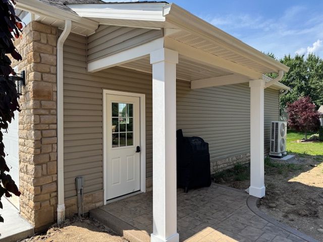 Covered side entrance with white door and columns on a beige house, beside a stone wall and concrete patio.