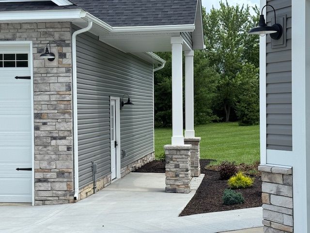 Covered concrete entryway with stone columns beside a gray house and landscaped yard.